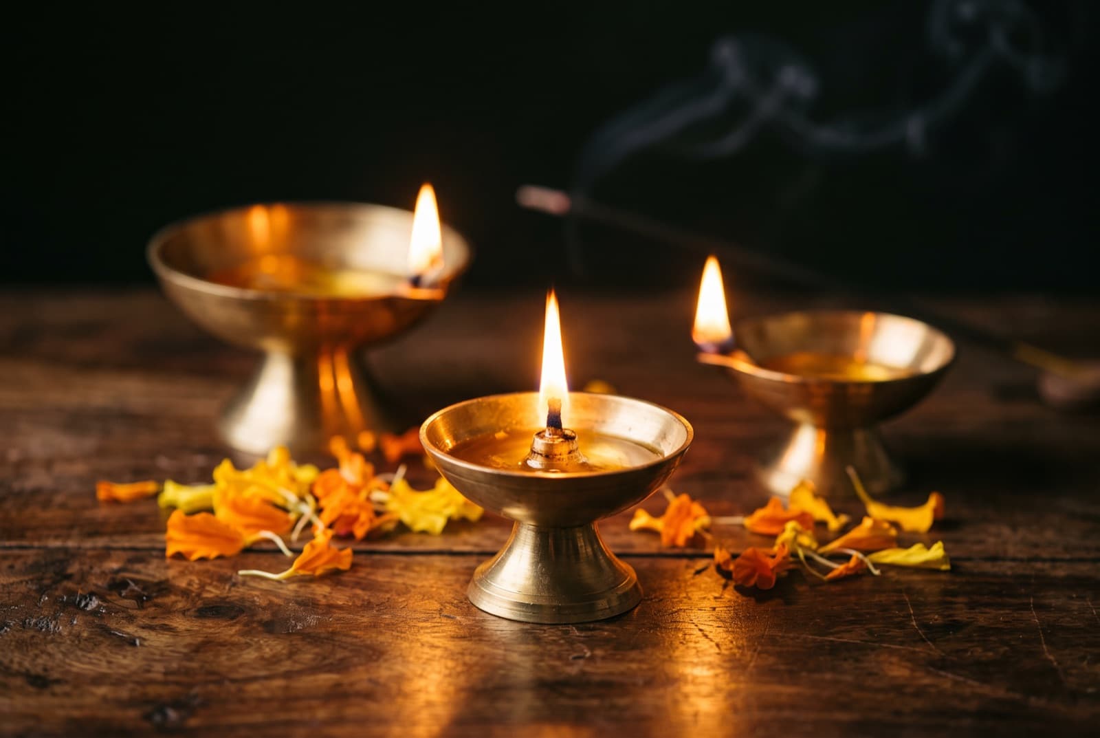 Three lit brass diyas on a dark wooden surface, incense smoke rising between them, marigold heads scattered around