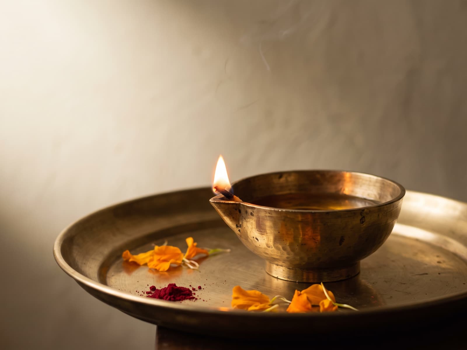 A lit brass diya resting on a brass thali with marigold petals and a pinch of kumkum, incense smoke drifting behind it